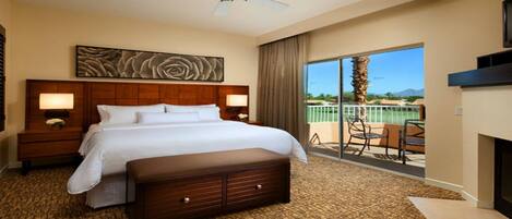 Habitación de hotel con una cama grande, cabecera de madera, dos lámparas de noche, un amplio ventanal con vistas a palmeras y un balcón con sillas.