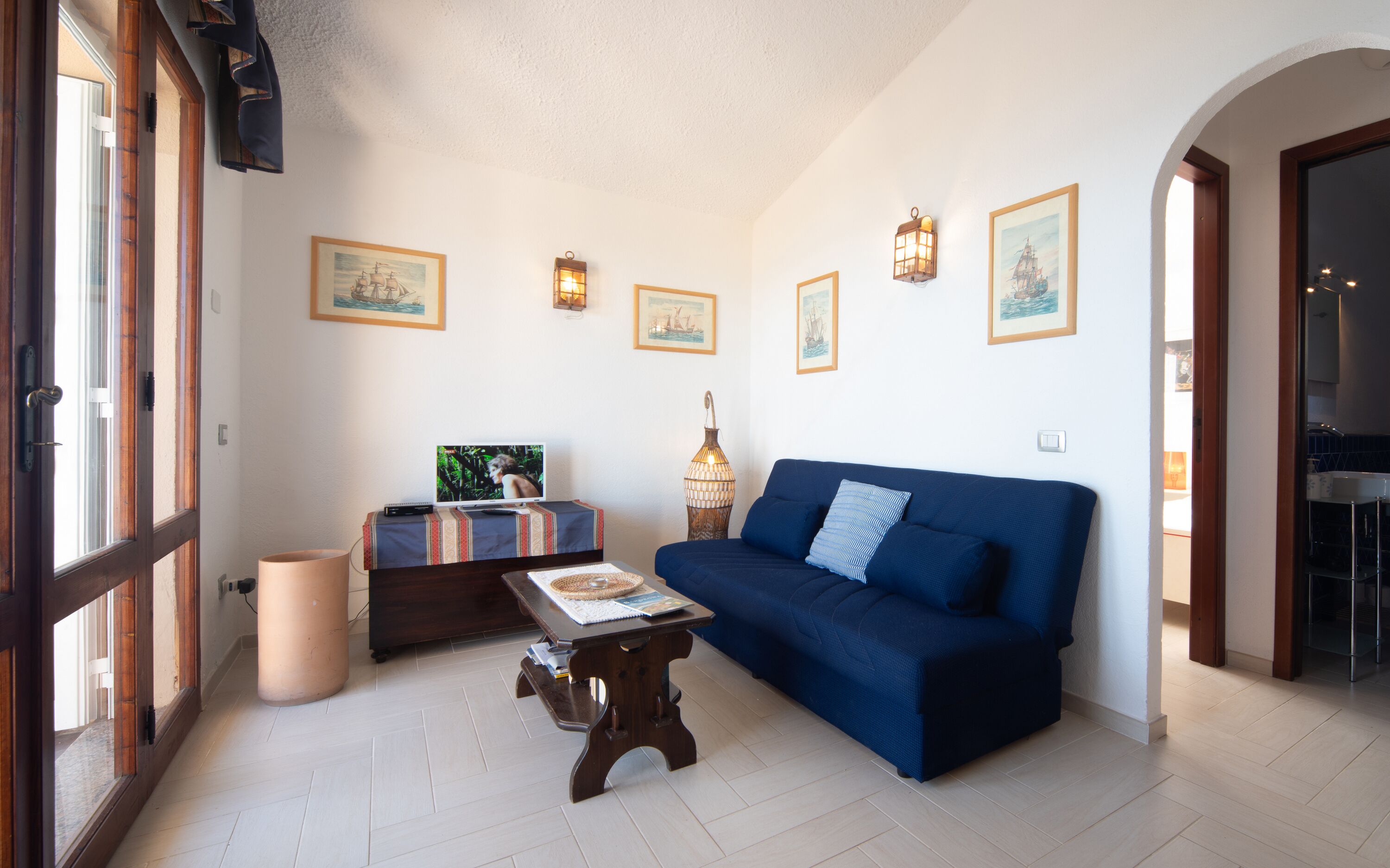 Een woonkamer met een blauwe bank, een kleine houten tafel en een televisie op een meubel. Er hangen afbeeldingen in lijsten aan de muur en er staat een vloerlamp.