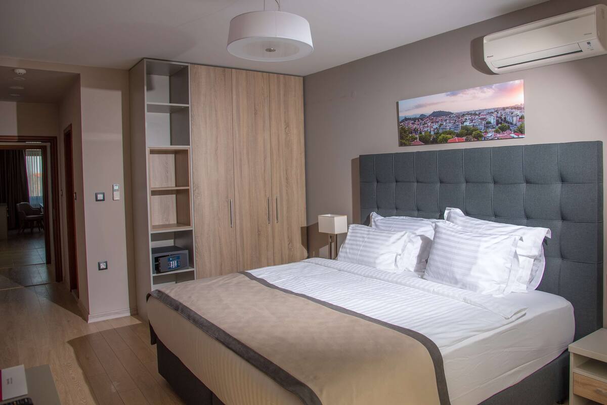 Een moderne slaapkamer met een groot bed, inbouwplanken, een grijs gestoffeerd hoofdeinde en een stadsgezicht schilderij aan de muur.