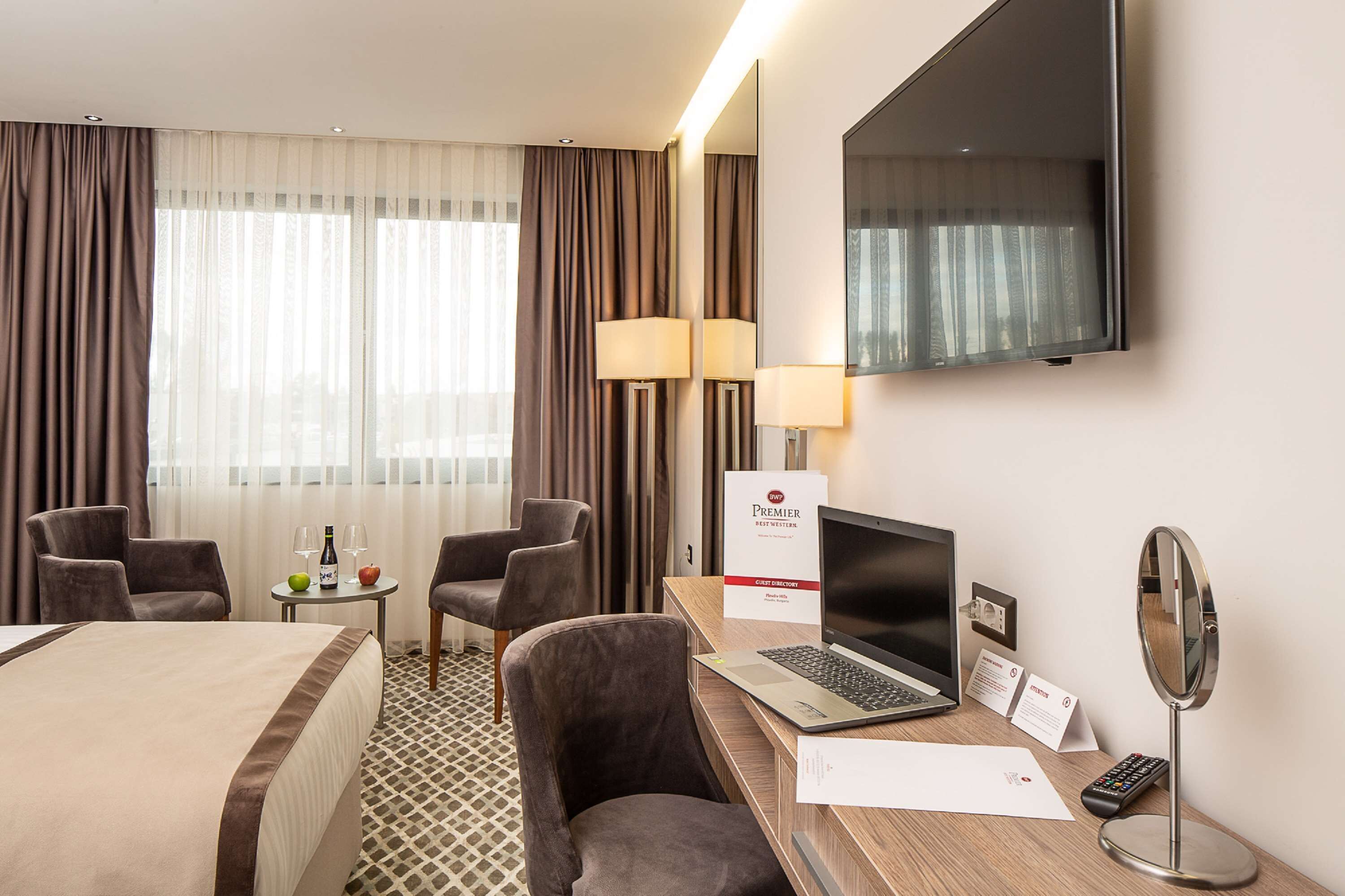 A hotel room with a bed, two armchairs, a desk with a laptop, a TV, a mirror, and a minibar.