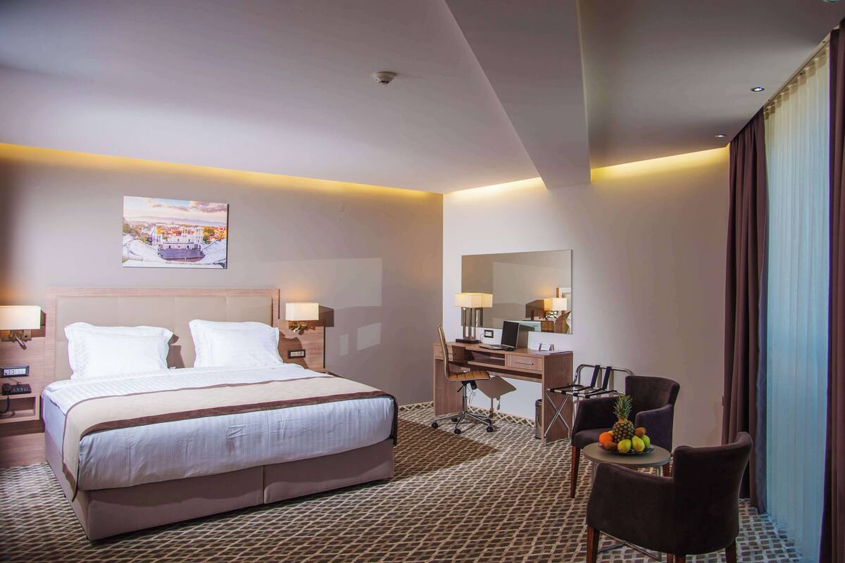 Een hotelkamer met een groot bed, een bureau met een computer, een stoel en een klein tafeltje met fruit.