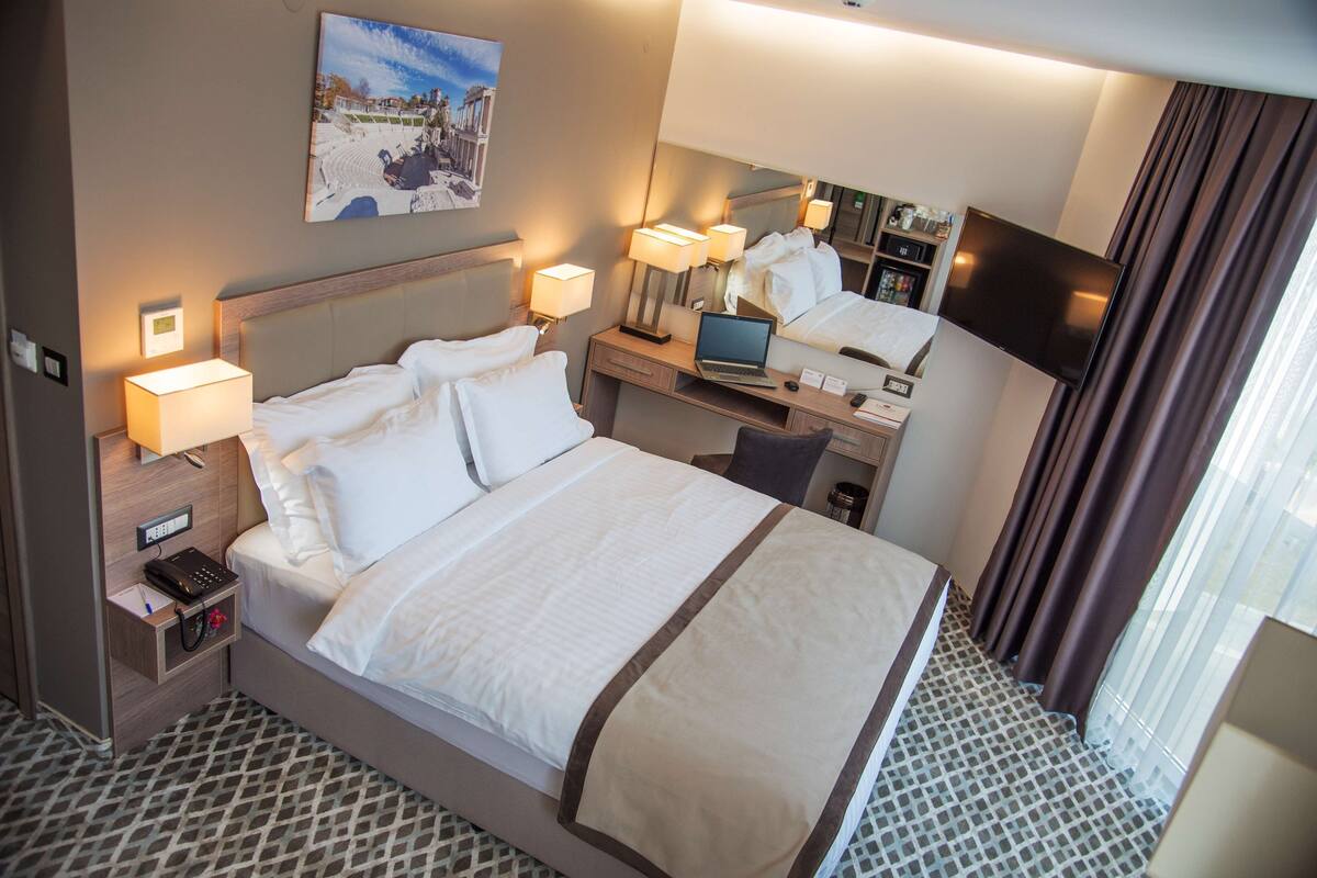 Een hotelkamer met een bed, nachtkastjes, een bureau met een laptop, een televisie en een raam met gordijnen.
