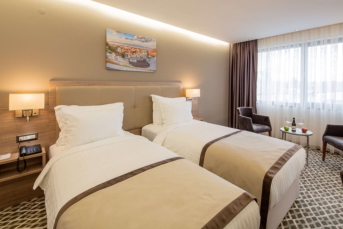 Hotelkamer met twee bedden, een bureau met lamp, een stoel, een tafeltje met fles en glazen, en een schilderij aan de muur.