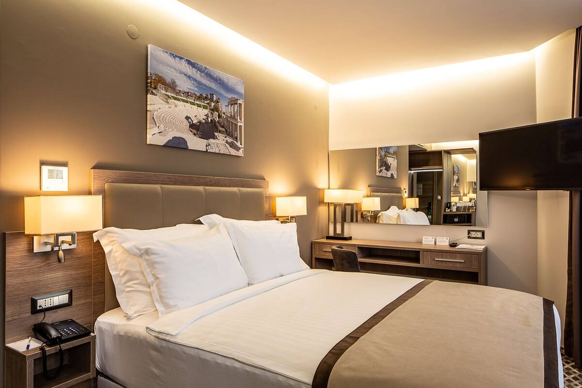 Een hotelkamer met een groot bed, nachtlampjes, een bureau met stoel, een flatscreen televisie en een schilderij aan de muur.