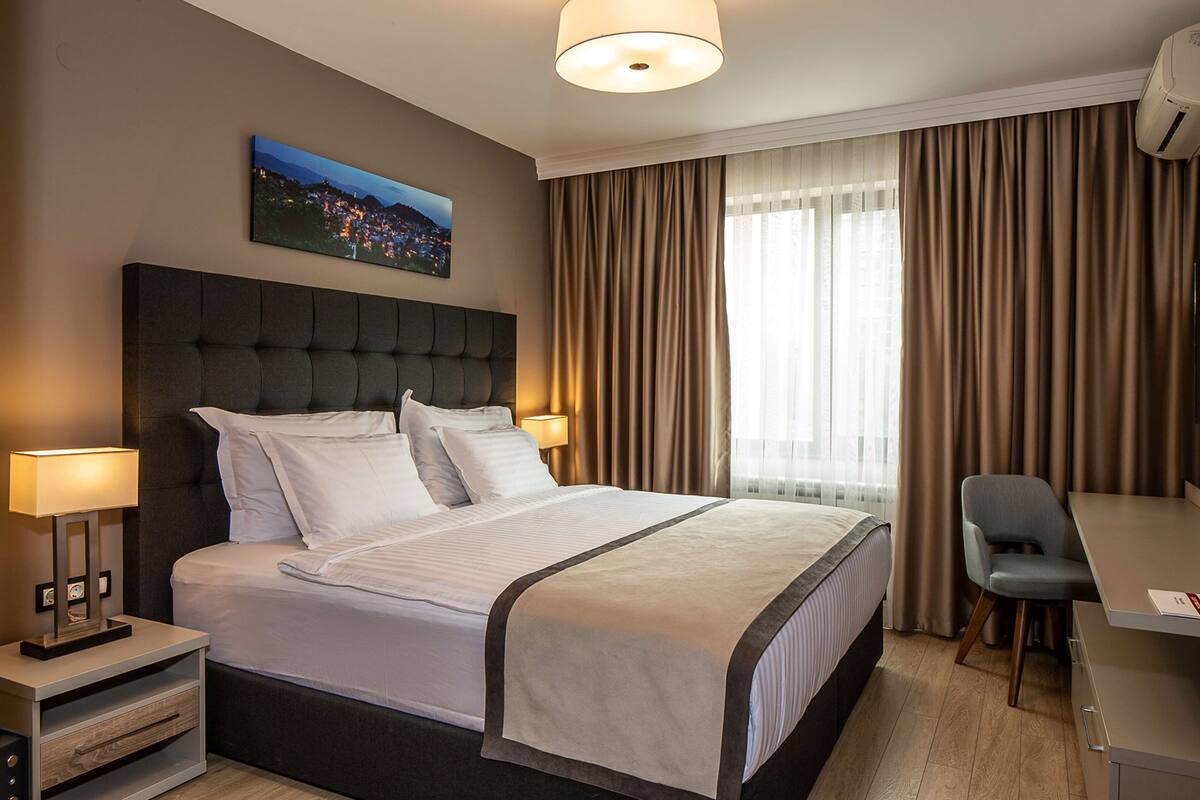 Een hotelkamer met een groot bed, een bureau en een stoel. Er hangt een foto aan de muur en er is een raam met gordijnen.