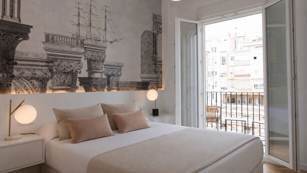 A bedroom with a large bed, a nightstand, a wall-mounted lamp, and a balcony with a view of buildings.