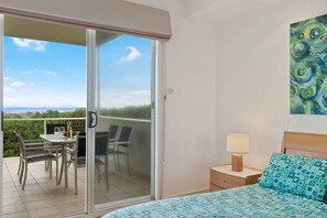 A bedroom with a bed, a nightstand, a lamp, a painting, and a balcony with a table and chairs. - Absolute Luxury Beachfront - Middleton (Middleton)