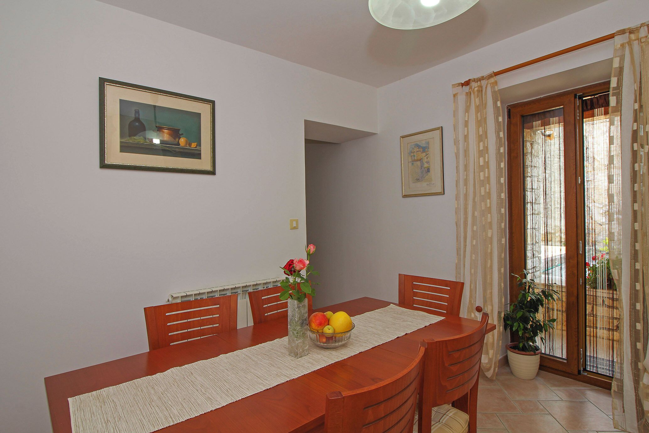 A dining area with a wooden table, chairs, a vase with flowers, and a bowl of fruit.