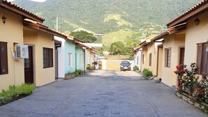 A paved street leading to a mountain view, flanked by buildings with tiled roofs and air conditioning units. - House in a gated community in São Francisco da Praia, São Sebastião/SP (Praia de São Francisco, São Sebastião)