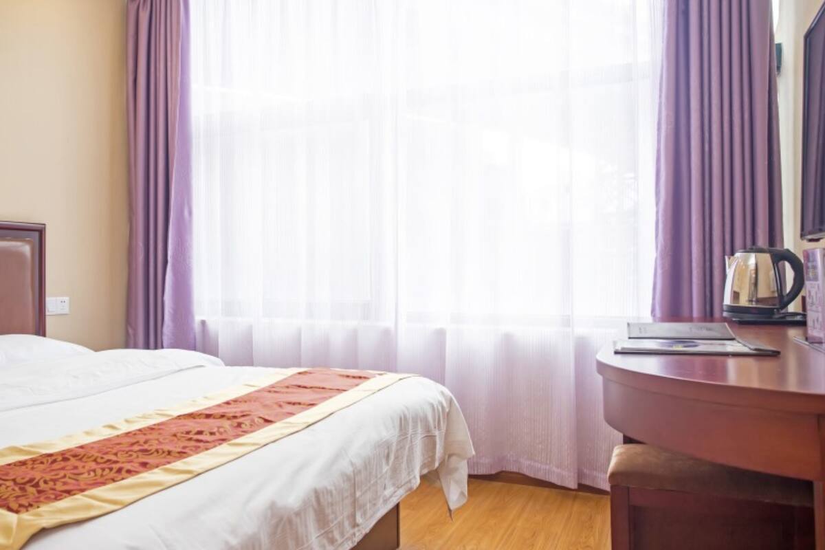 A hotel room with a bed, a wooden desk, a kettle, and a window with curtains.