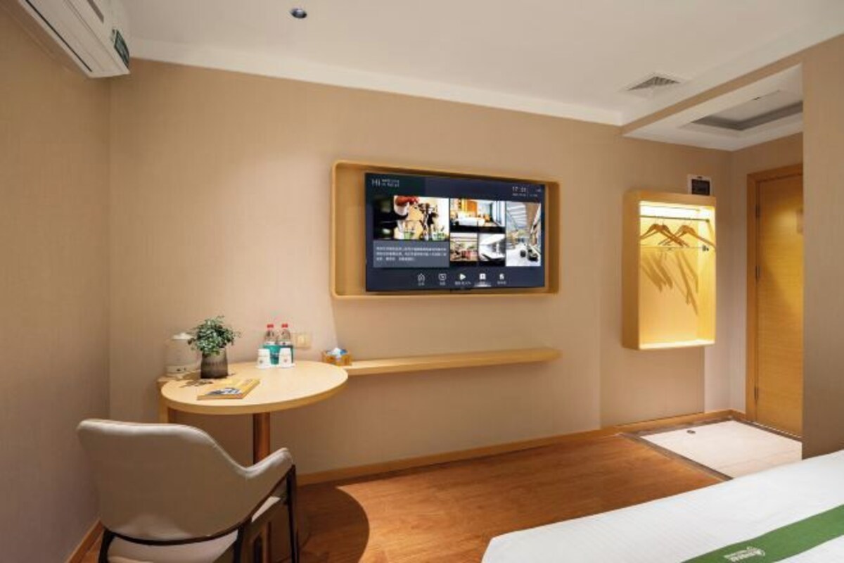 Une chambre d’hôtel avec un lit, un bureau, une chaise, un téléviseur fixé au mur et une petite table sur laquelle se trouvent une plante et des bouteilles.