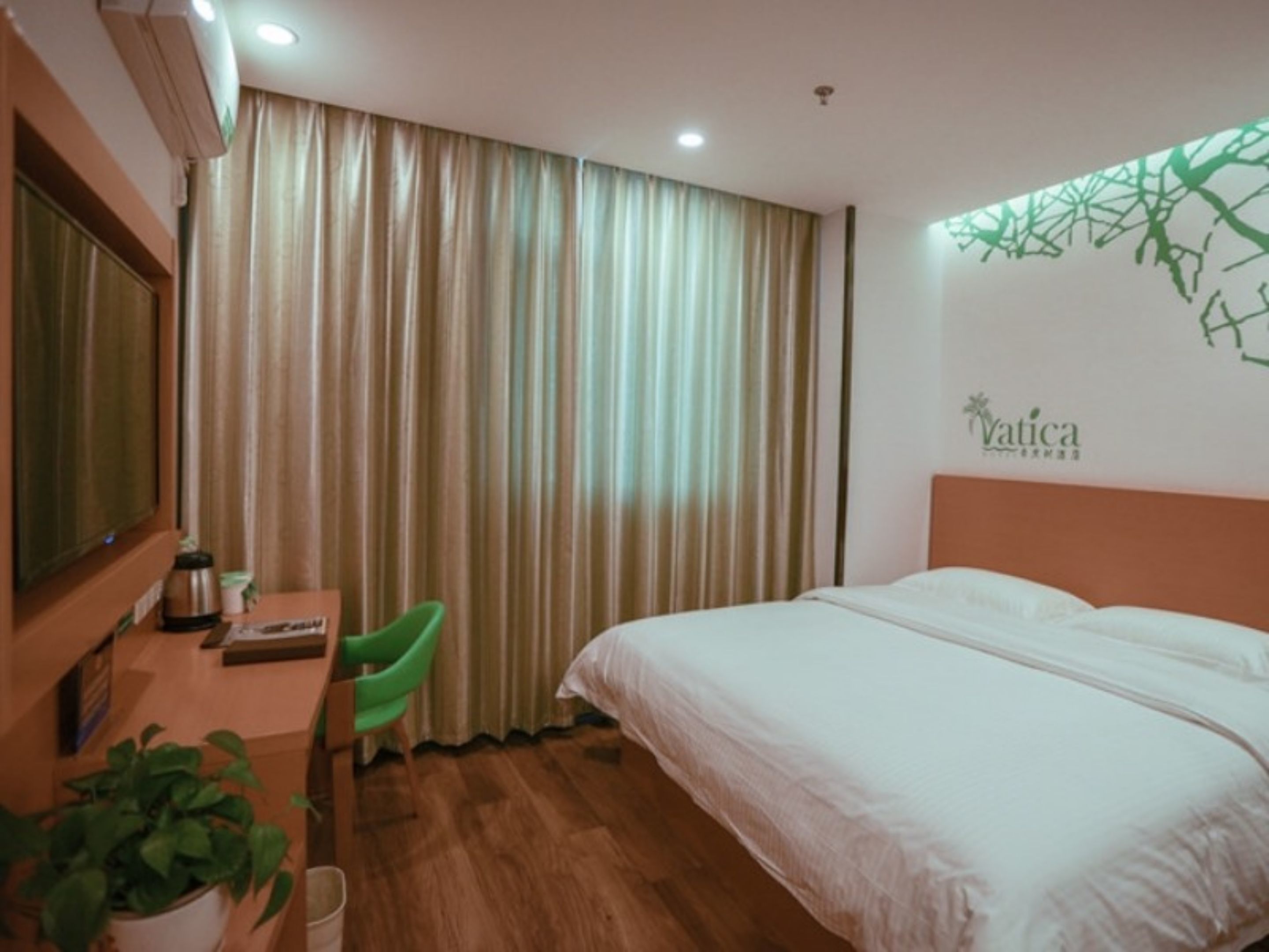 A hotel room with a bed, a desk with a chair, a potted plant, and a wall with a logo.