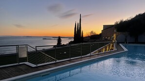 Ein Schwimmbecken mit Holzterrasse und Metallgeländer, mit Blick auf ein Gewässer, in der Ferne Land und Bäume bei Sonnenuntergang.