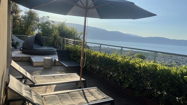 Eine Terrasse mit Sitzgelegenheit, einem großen Sonnenschirm und Blick auf das Meer und die Berge.