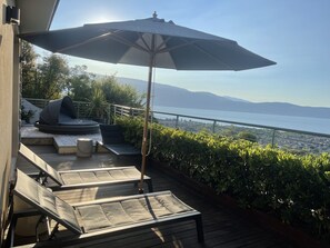 A deck with outdoor seating, a large umbrella, and a view of the sea and mountains.