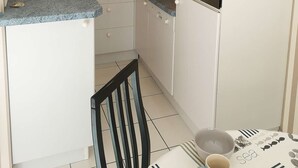 A compact kitchen with white cabinets, a built-in oven, a kettle, and a dining area with a chair and table. - Arcachon – One-bedroom flat with sea views in a quiet location (ARCACHON)
