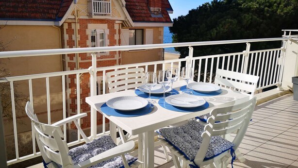 A balcony with a table set for four, white chairs with patterned cushions, and a view of a building with a red roof. - Arcachon – One-bedroom flat with sea views in a quiet location (ARCACHON)