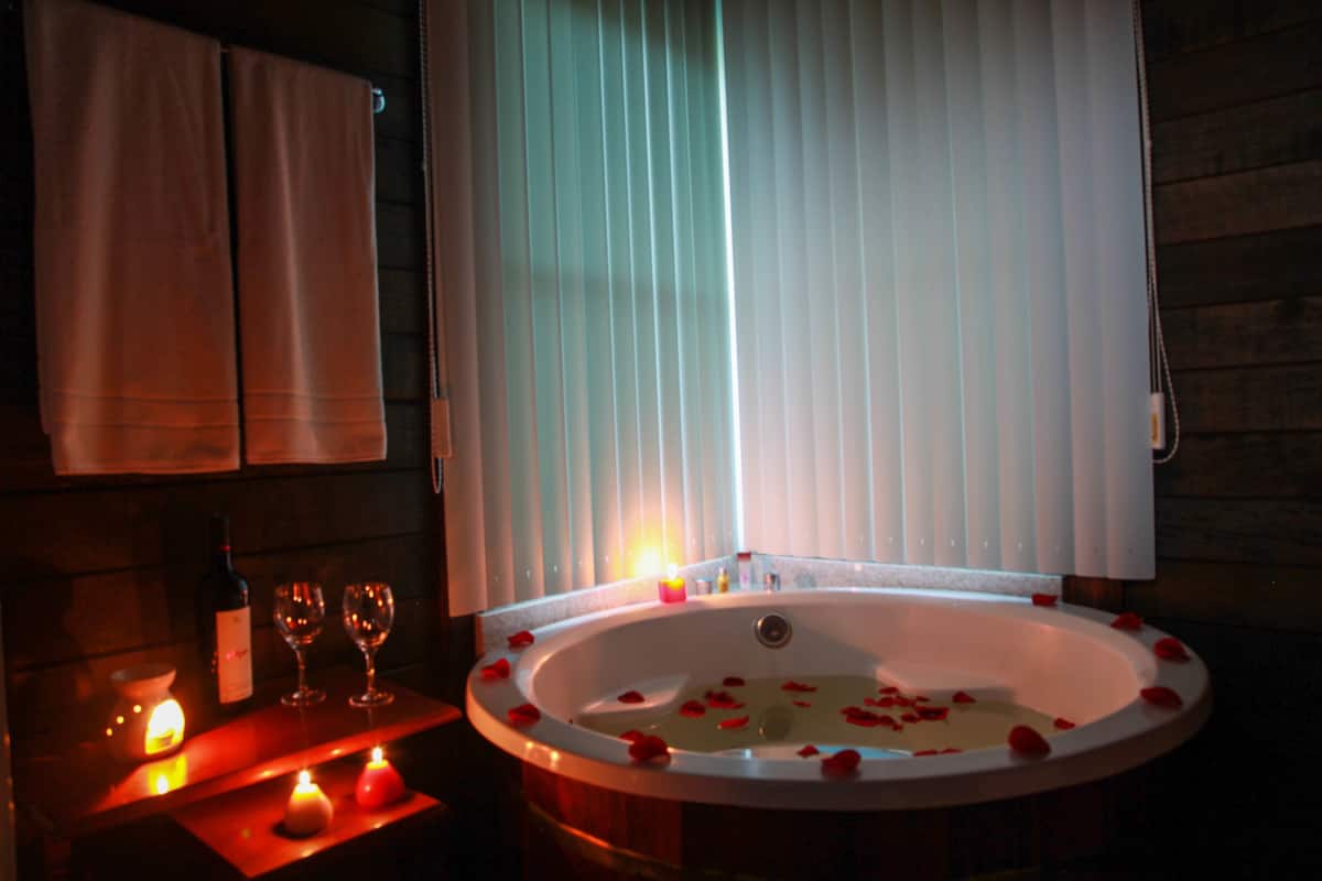 A dimly lit room with a bathtub filled with rose petals, candles, and wine glasses on a wooden table.