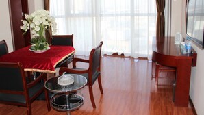 A hotel room with a dining area featuring a round table with a red tablecloth, four chairs, a vase of white flowers, and a small glass table with a candle. There is a flat-screen TV on a wooden stand and a large window with curtains.