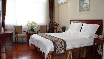 A hotel room with a large bed, a desk, and a chair. There is a window with curtains, an air conditioning unit, and a painting on the wall.