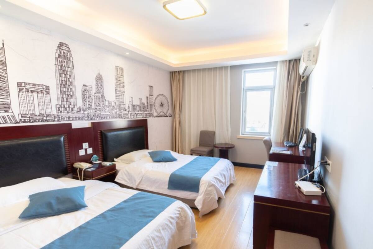 A hotel room with two beds, a desk, a chair, a small table, a window with curtains, and a cityscape mural on the wall.