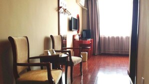 A hotel room with a wooden floor, a TV mounted on the wall, a desk with a chair, and a window with curtains.