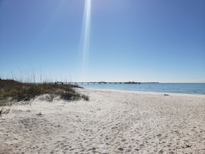 Ein Strand mit einem Steg, der ins Wasser führt, feinem Sand und vereinzelter Vegetation.