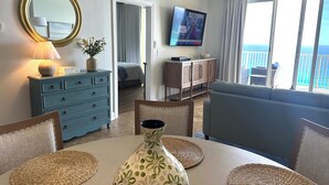 A living room with a TV, a sofa, a dining table with placemats, a dresser, and a view of the ocean. - Emerald Isle 1708 Just Remodeled! Stunning! Front Row Beach Chairs Included! (Panama City Beach)