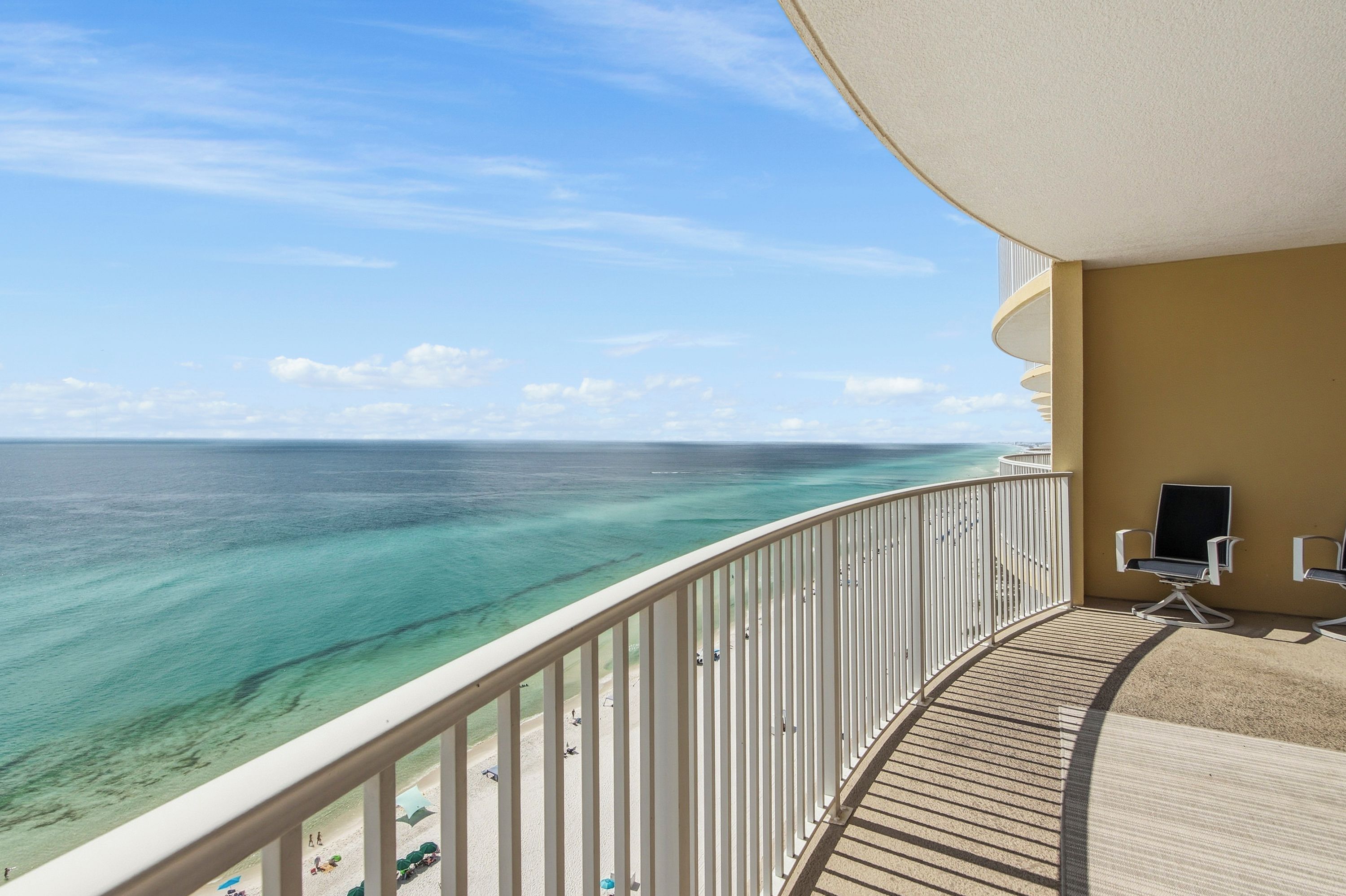 A balcony with a view of the ocean, a beach, and a lounge area with chairs and a TV.