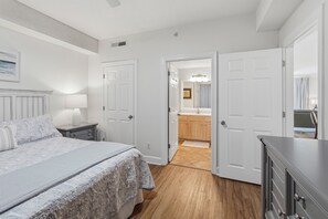 Quarto com cama, mesa de cabeceira, luminária e porta que dá para um banheiro com pia e espelho.