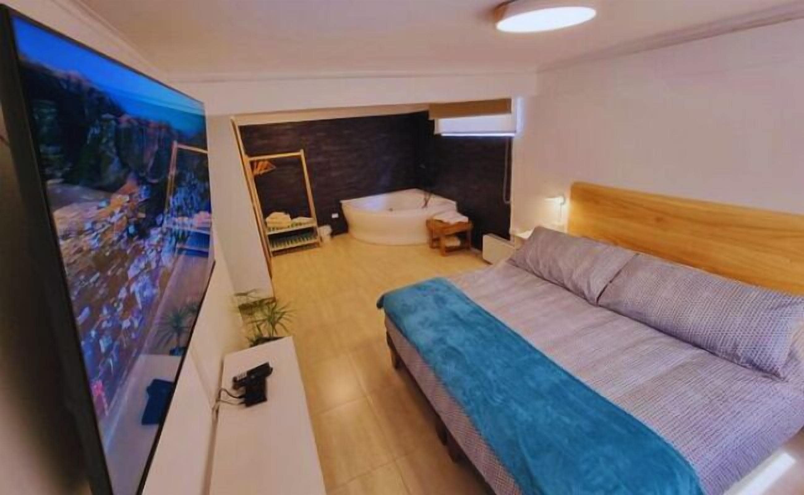 A modern bedroom with a bed, a small bedside table, a wall-mounted TV, and a bathtub in the background.