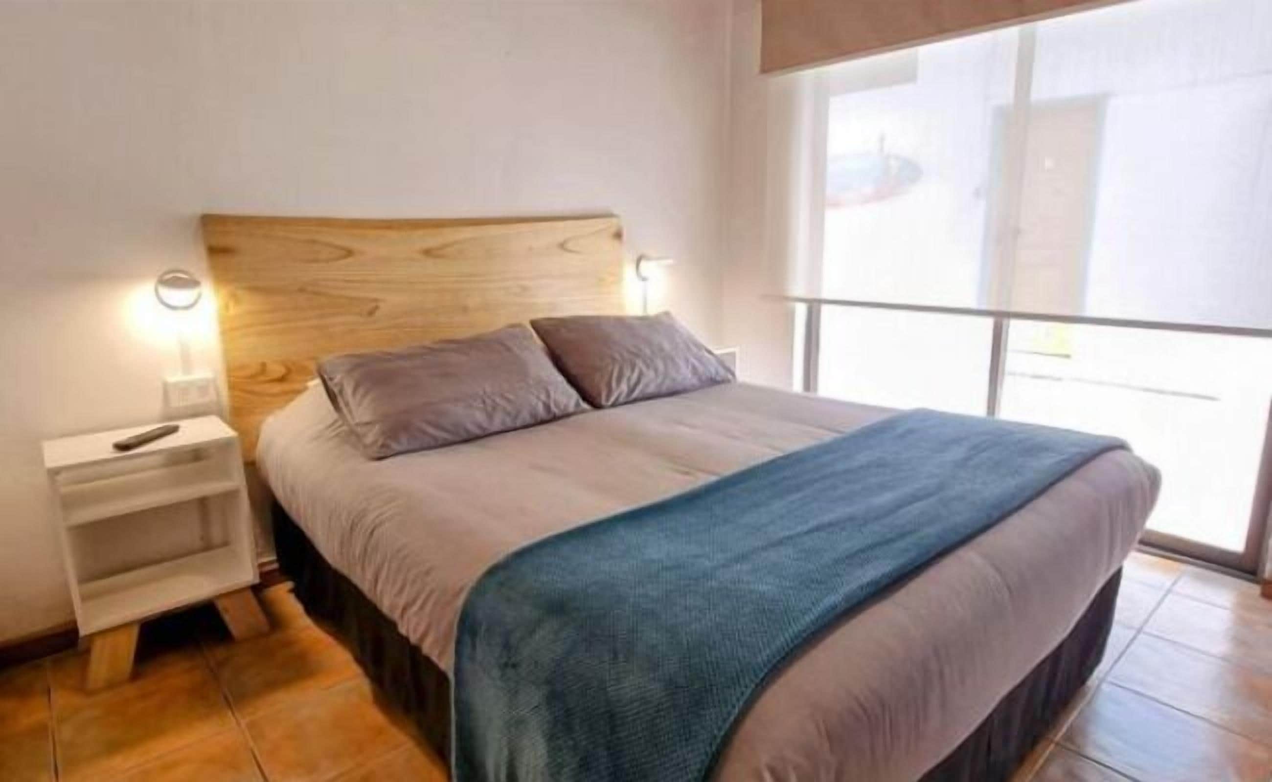 Een slaapkamer met een houten hoofdbord, een bed met een blauwe deken, twee nachtkastjes met lampen en een groot raam.