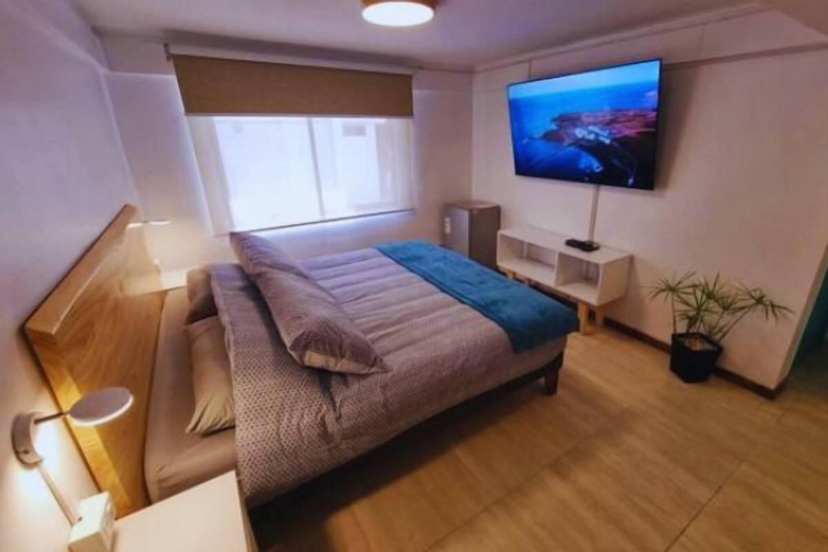 Un dormitorio con cama, televisor en la pared, mesita de noche y ventana.