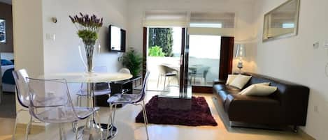 A modern living room with a glass dining table, clear plastic chairs, a dark rug, a brown sofa, and a TV on the wall.