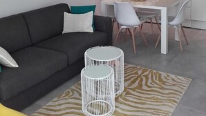 A modern living room with a grey sofa, white dining table, and chairs. There is a kitchen area with white cabinets and a built-in oven. Two white, cylindrical side tables are placed in front of the sofa. - Villa King (Dubrovnik)