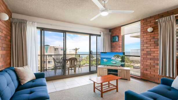 Ein Wohnzimmer mit Fernseher, zwei blauen Sofas, einem Holztisch und einem Balkon mit Meerblick.