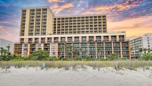 Ein mehrstöckiges Hotel mit flachem Dach und Balkonen, gelegen in Strandnähe mit Sand und Vegetation.