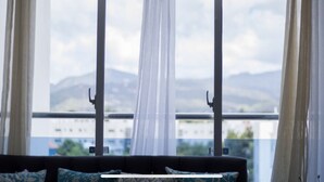 A room with a bed, a window with sheer curtains, and a view of mountains and buildings.