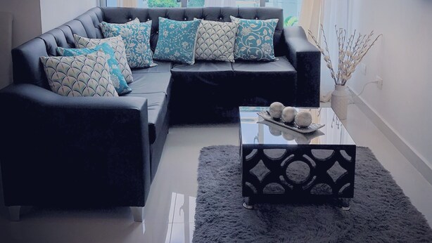 A modern living room with a black leather sofa, patterned cushions, a glass coffee table, and a view of a cityscape through large windows.