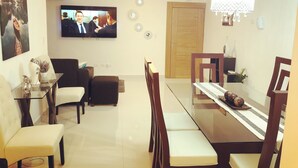 A modern living room with a flat-screen TV, a chandelier, and a dining area with a glass table and chairs.