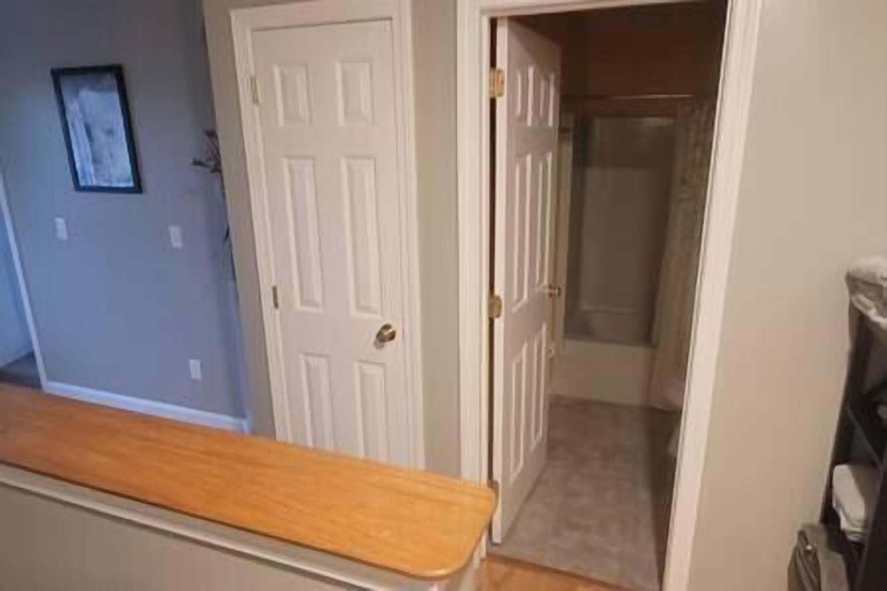 A hallway with a wooden bench, a white door, and a view into a room with a carpeted floor.
