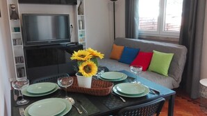 Un comedor con una mesa dispuesta para cuatro personas, un televisor en la parte superior y un sofá con cojines de colores al fondo.