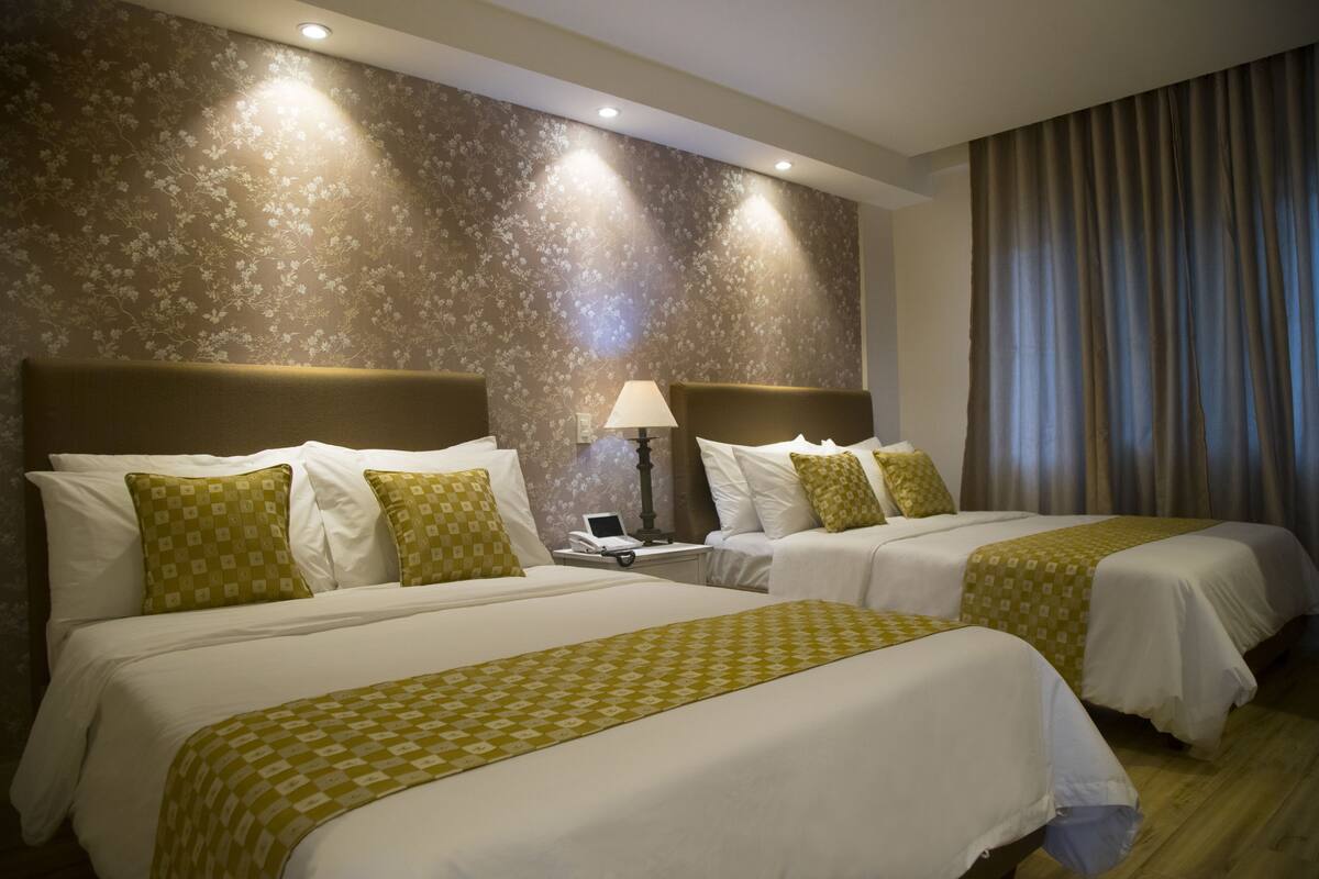 A hotel room with two beds, a nightstand with a lamp, and a floral-patterned wall.