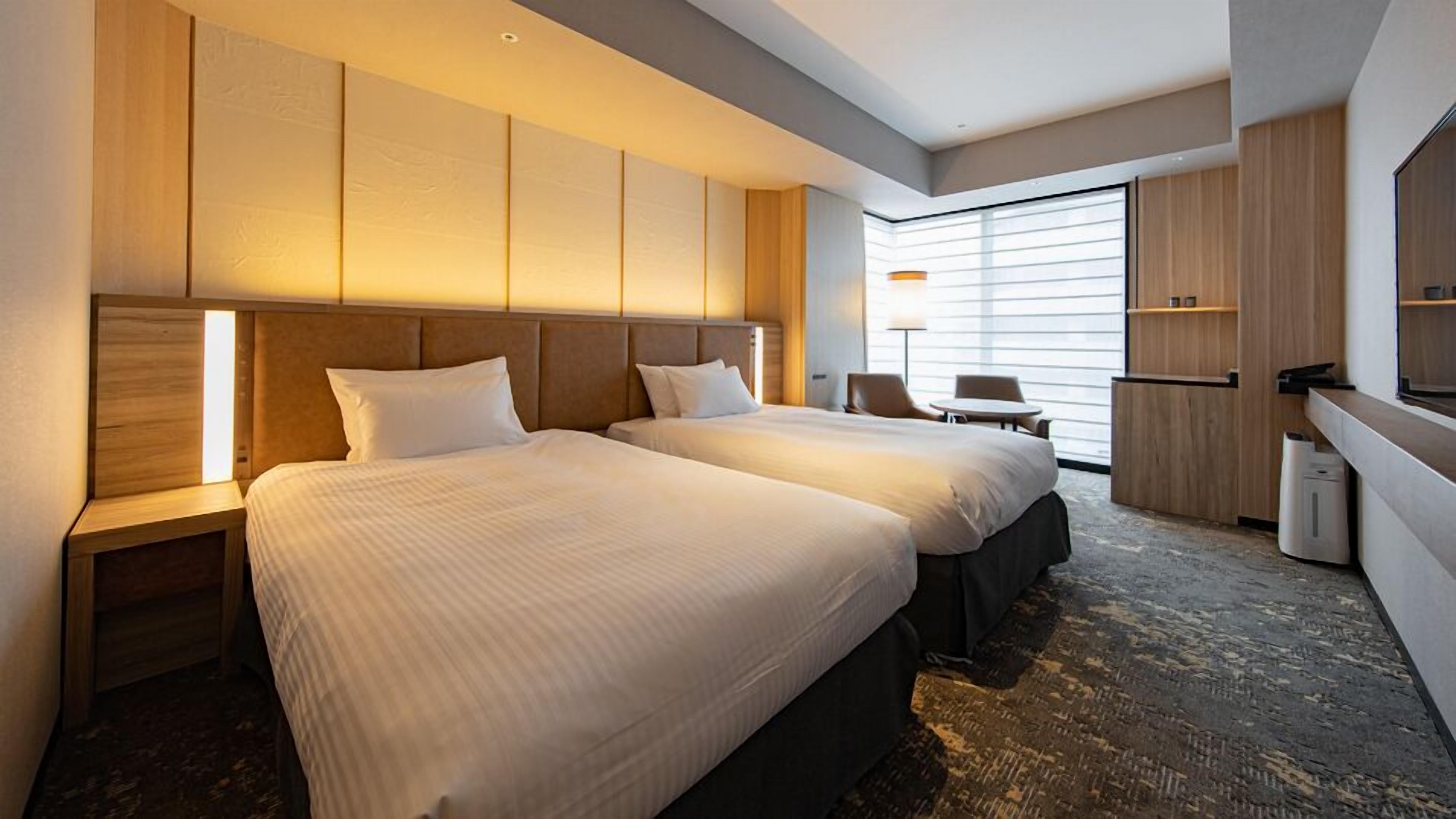 A hotel room with two beds, a desk, a chair, and a large window.