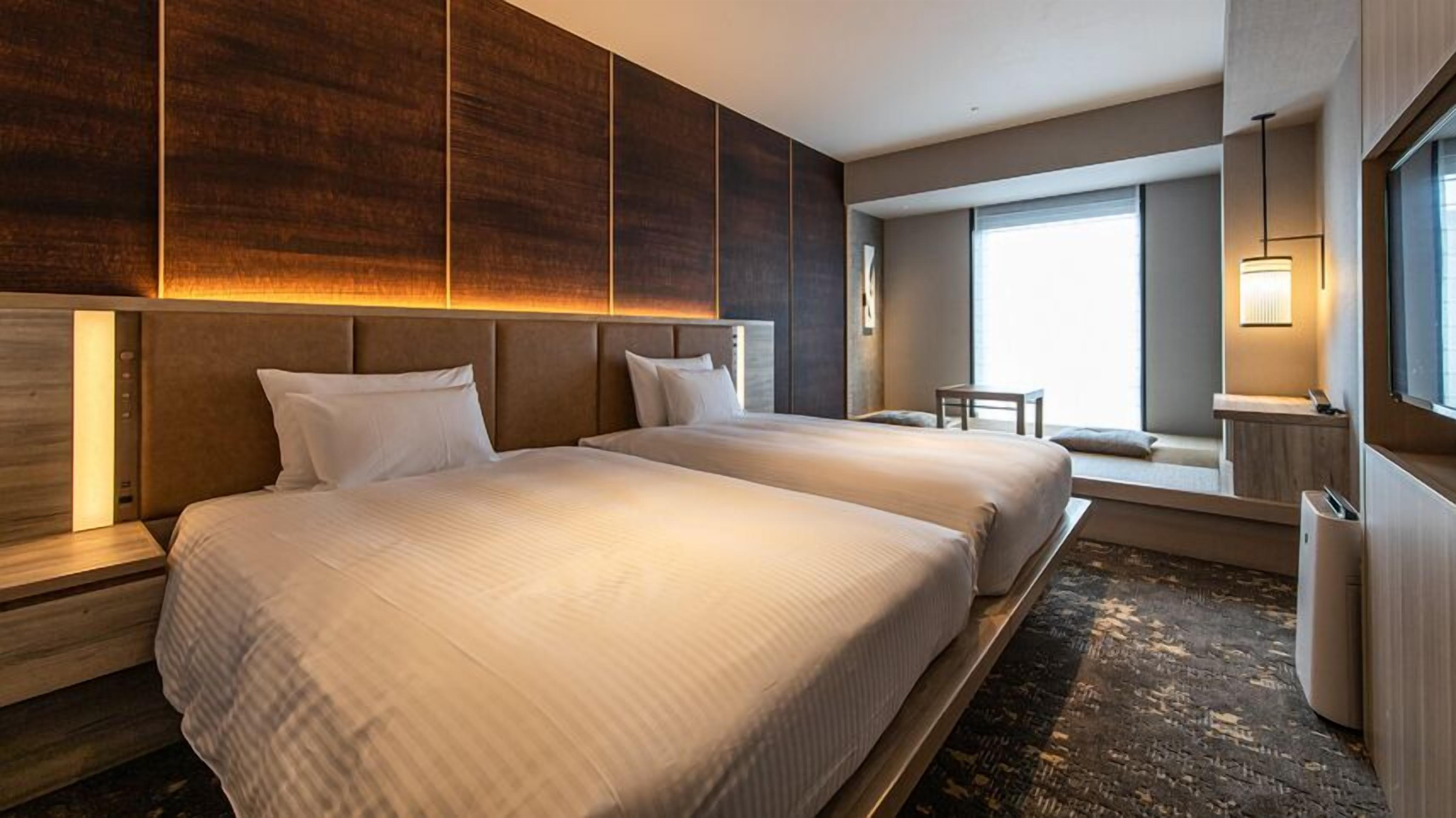 A hotel room with two beds, a desk, a chair, and a large window.