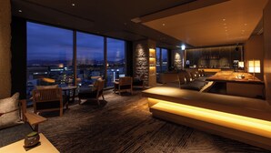 A modern living room with a large window offering a city view, a cozy seating area, and a kitchen area. - THE BLOSSOM HAKATA Premier (Fukuoka)