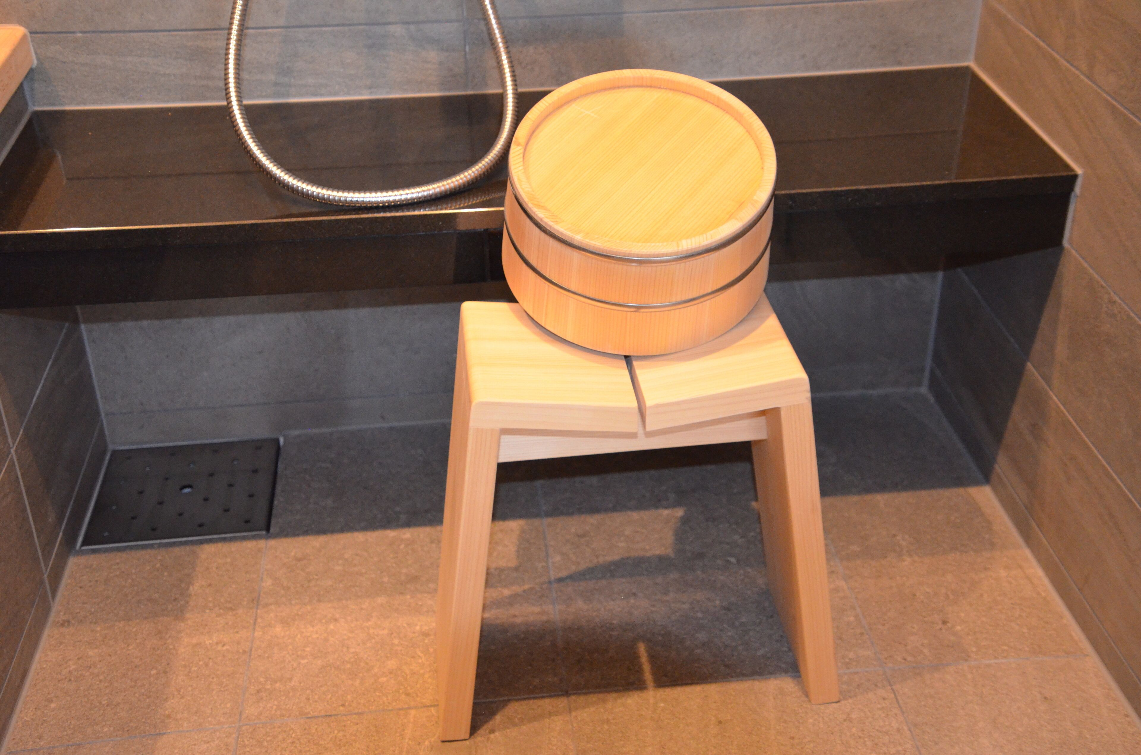 A wooden stool with a round seat in a bathroom with dark tiled walls and a black countertop.