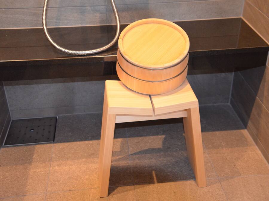 A wooden stool with a round seat in a bathroom with dark tiled walls and a black countertop.