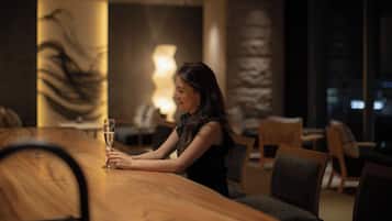 A woman sitting at a wooden table in a dimly lit room with a large abstract painting on the wall and a lamp in the background.