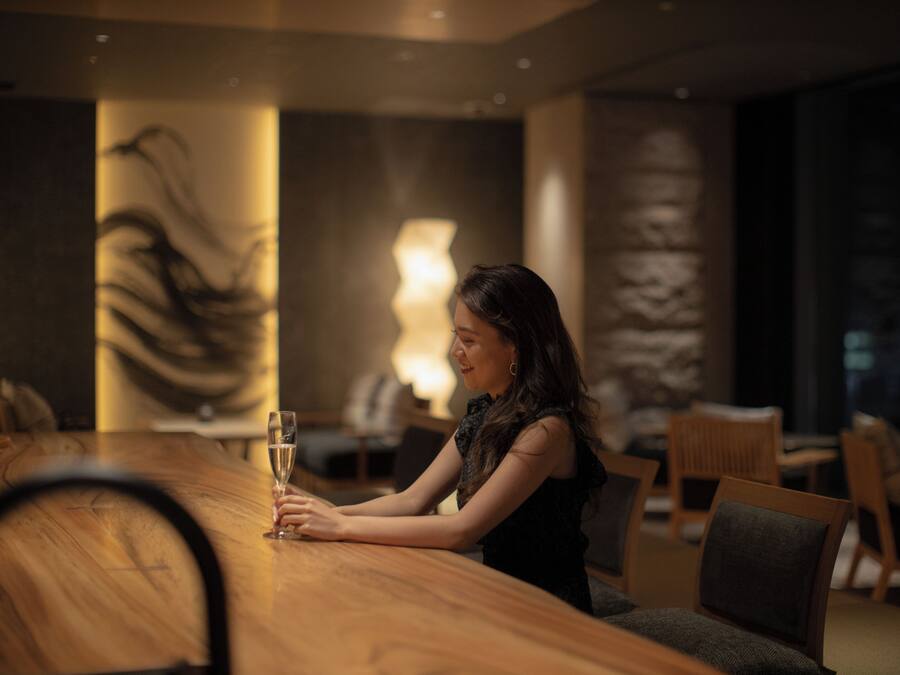 A woman sitting at a wooden table in a dimly lit room with a large abstract painting on the wall and a lamp in the background.